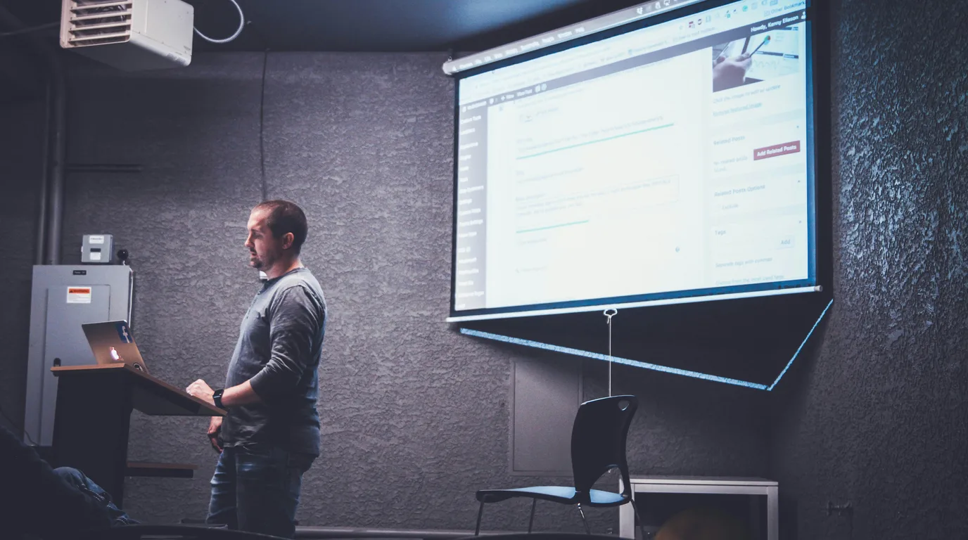 A person at a lectern in a dark room, facing a projector screen showing a web publishing dashboard