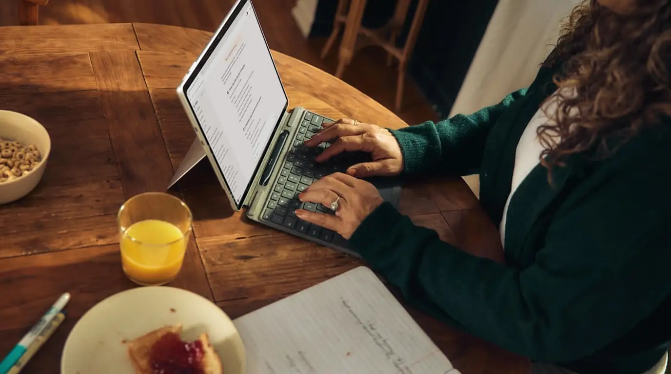 Overhead: a person in a green cardigan typing on a tablet keyboard at a wooden table, cereal and juice beside