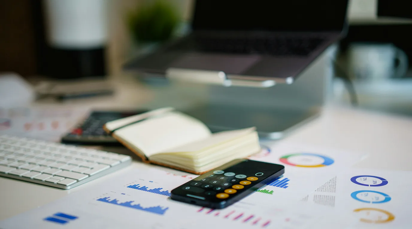 A desk with printed bar and donut charts, a calculator phone, an open notebook, keyboard and laptop